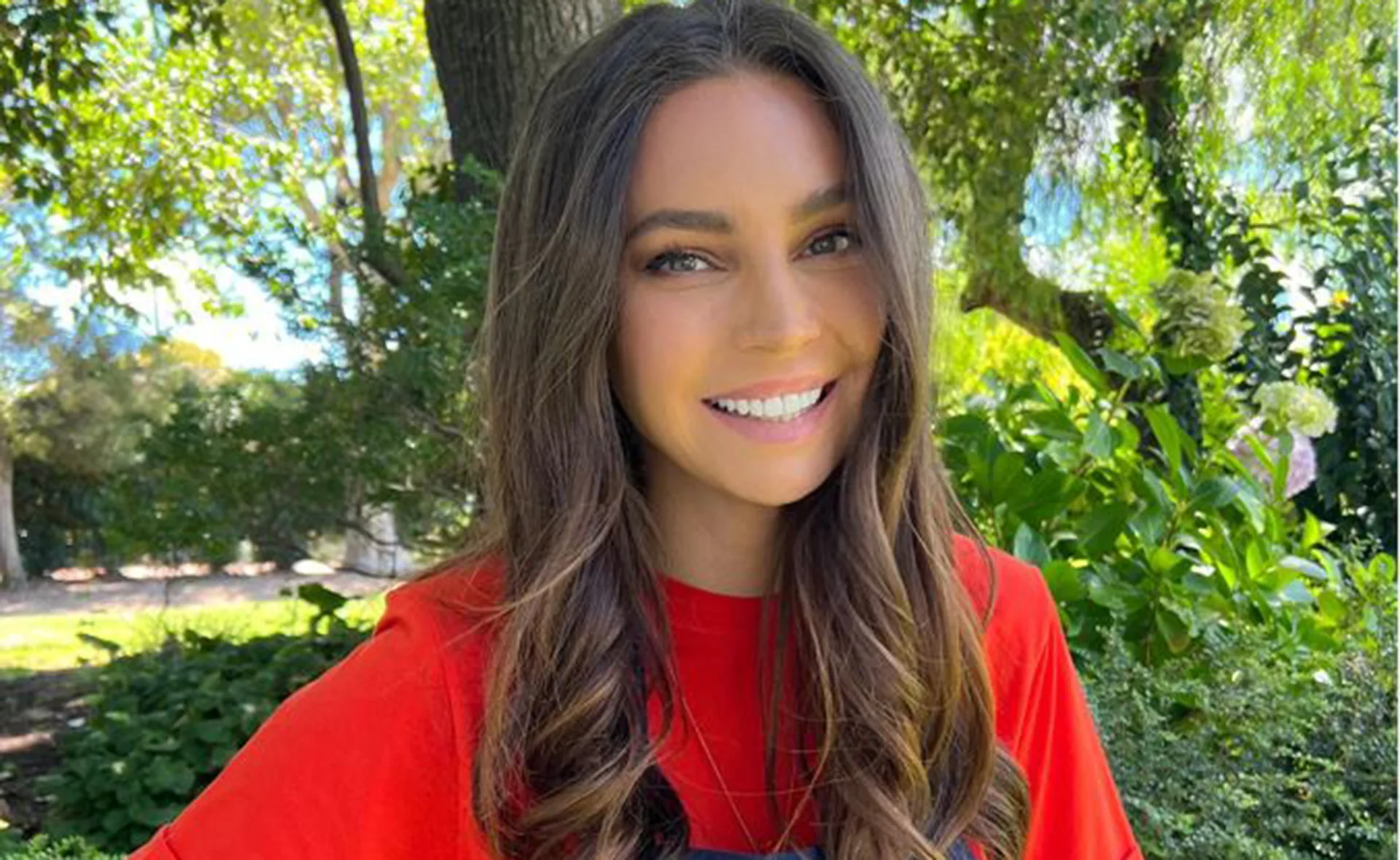 Person smiling outdoors surrounded by green foliage and trees, wearing a red shirt.