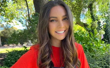 Person smiling outdoors surrounded by green foliage and trees, wearing a red shirt.