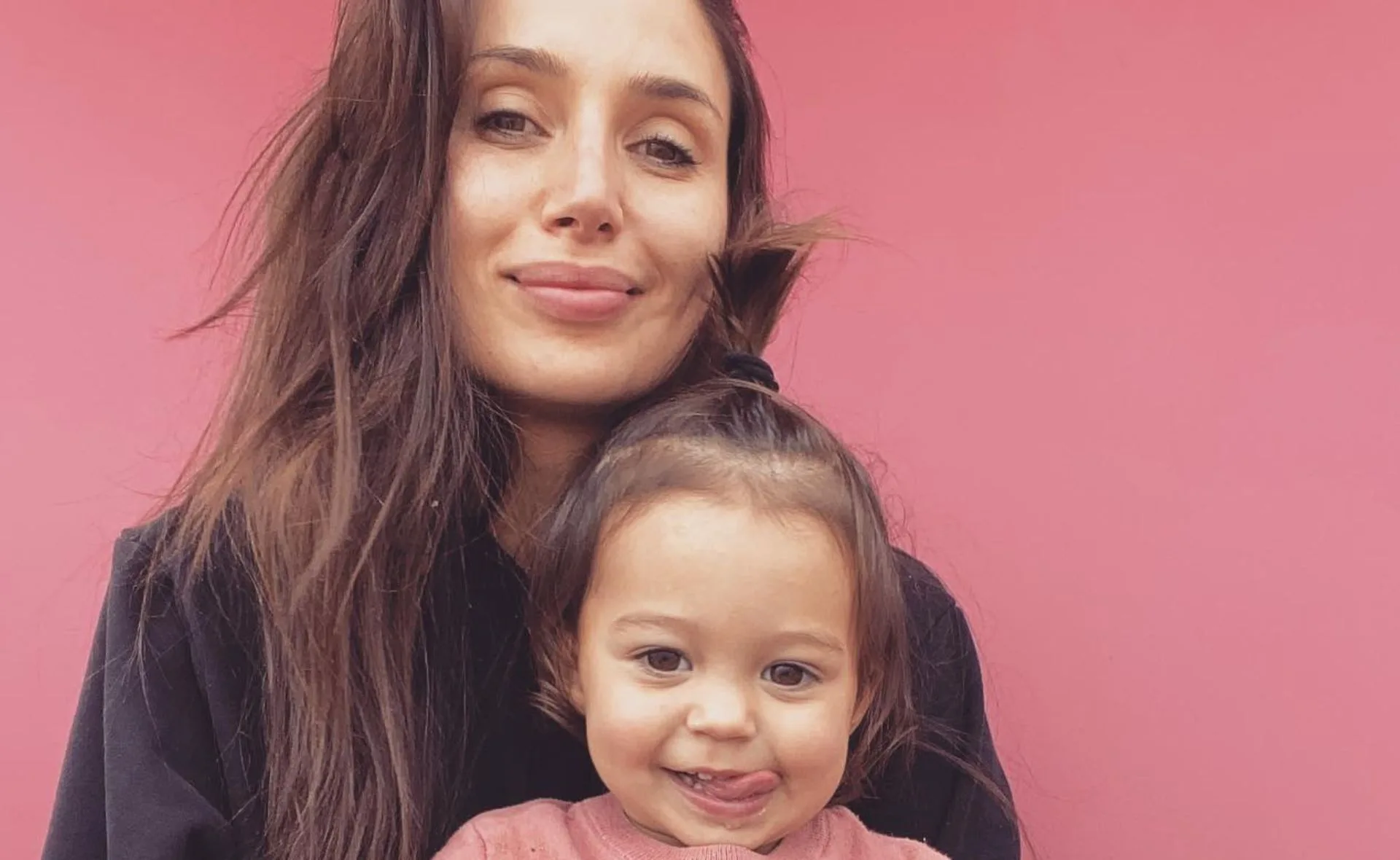 Woman with long brown hair and a toddler smiling in front of a pink background.