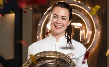 Chef in a white coat holding a trophy, smiling with confetti falling around her at a competition.