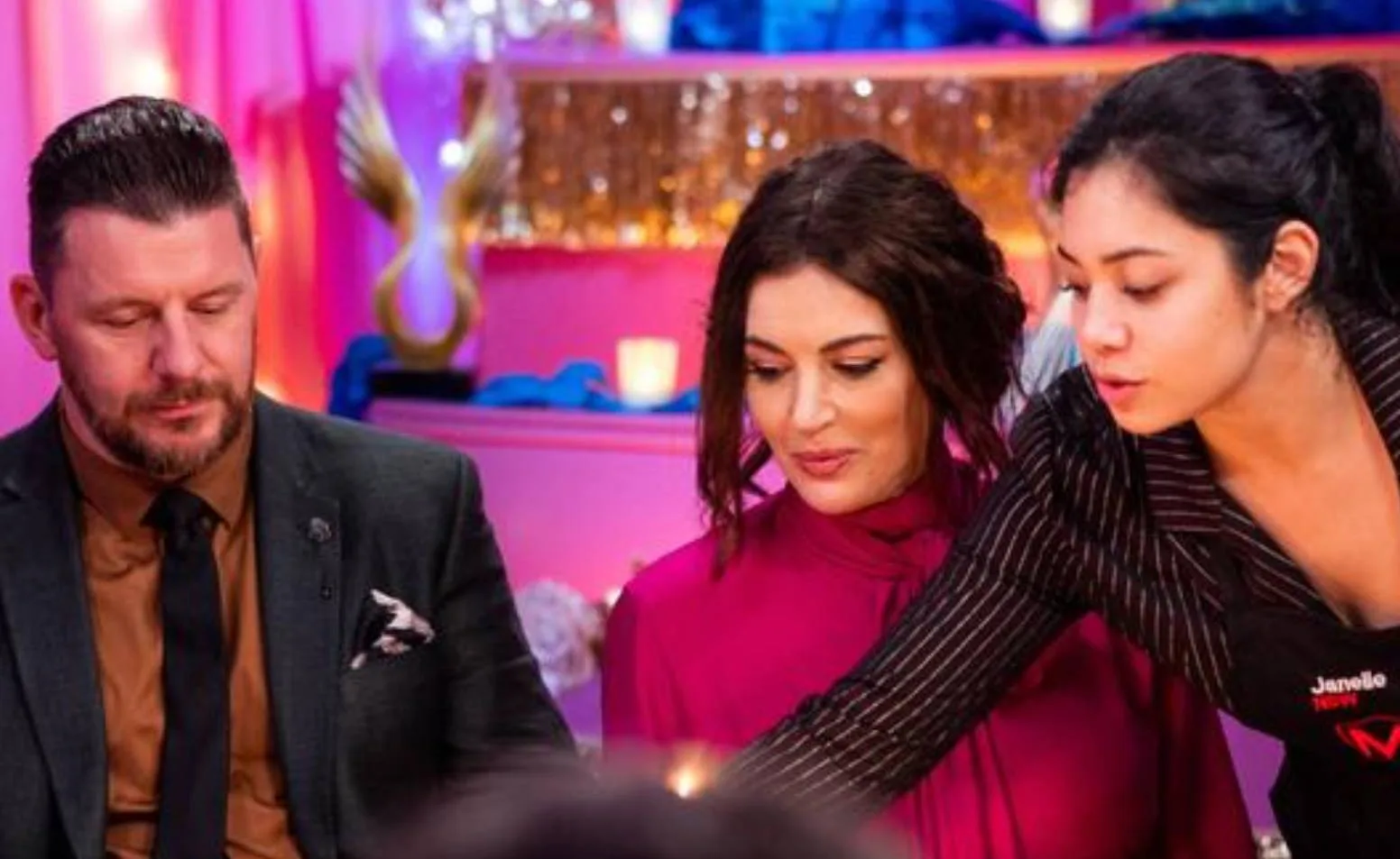 Three people in a colorful room, one serving food, with pink and blue decor in the background.