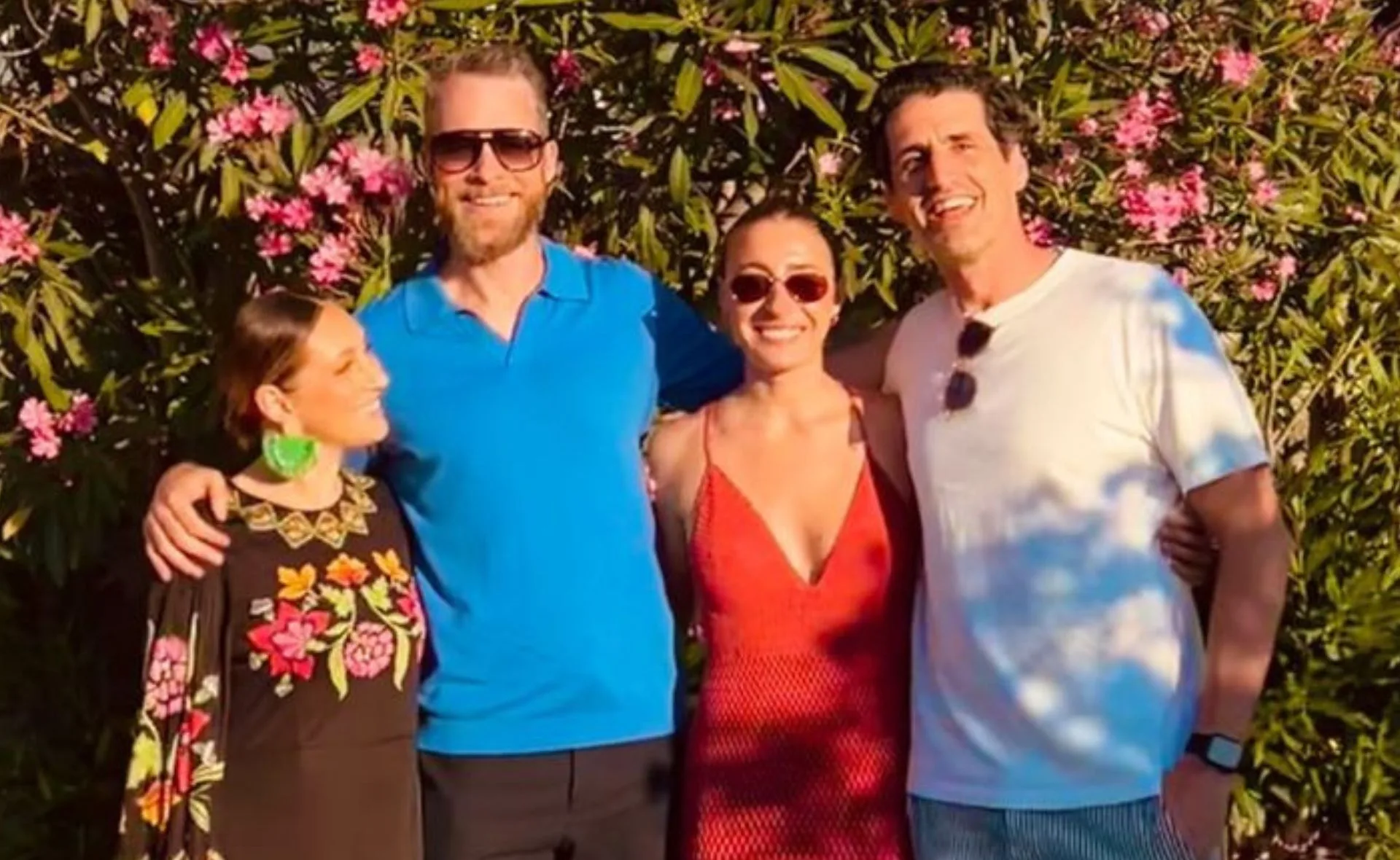 A group of four friends smiling in front of flowering bushes with colorful outfits and sunglasses.