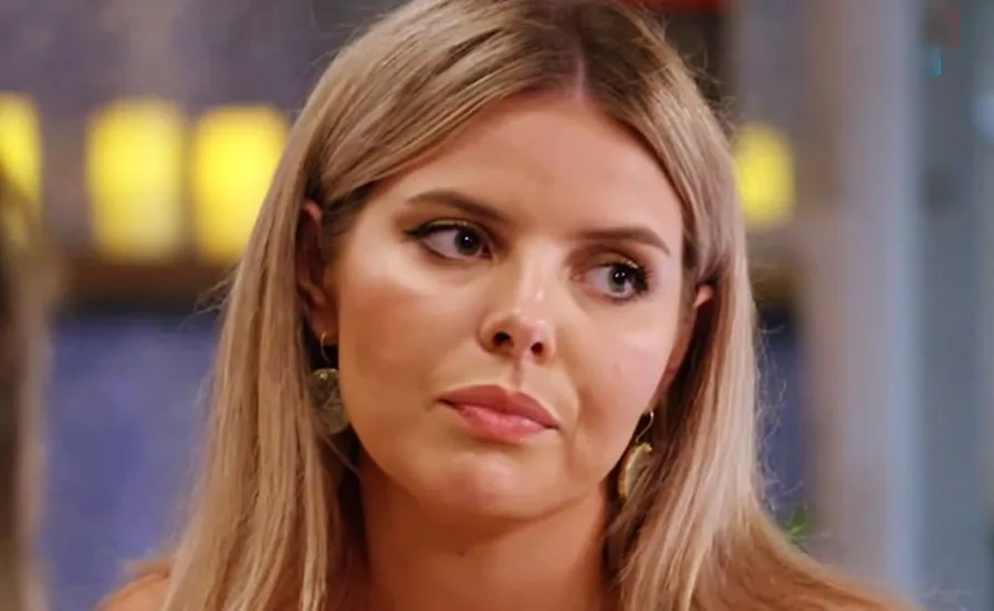 A person with long hair looking to the side with a neutral expression, wearing earrings; blurred background.