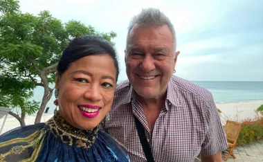 Smiling man and woman taking a selfie on a beach with greenery and ocean in the background.