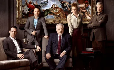 The main cast of "Succession" in formal attire, standing and seated in a luxurious room with a large painting behind them.