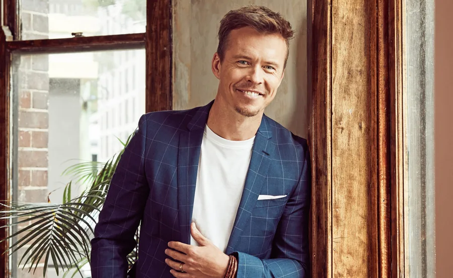 Man in a blue checkered suit jacket smiling while standing by a window with a plant in the background.