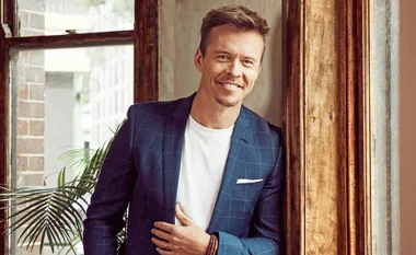 Man in a blue checkered suit jacket smiling while standing by a window with a plant in the background.