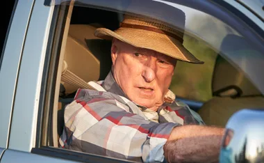 Elderly man in a checked shirt and straw hat sitting in a car, looking to the side with a serious expression.