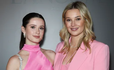 Two women smiling, wearing pink outfits, posing for a photo against a neutral background.
