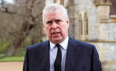 Man in a suit and tie outside a stone building, with trees and blurred background.