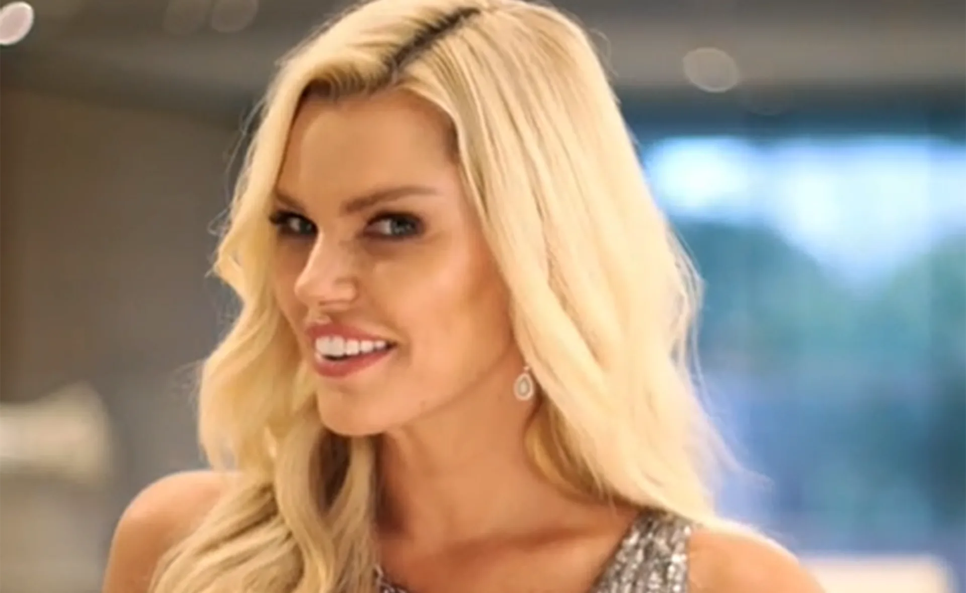 Blonde woman smiling, wearing a sparkly dress and earrings, with a blurred indoor background.