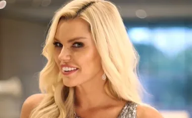 Blonde woman smiling, wearing a sparkly dress and earrings, with a blurred indoor background.