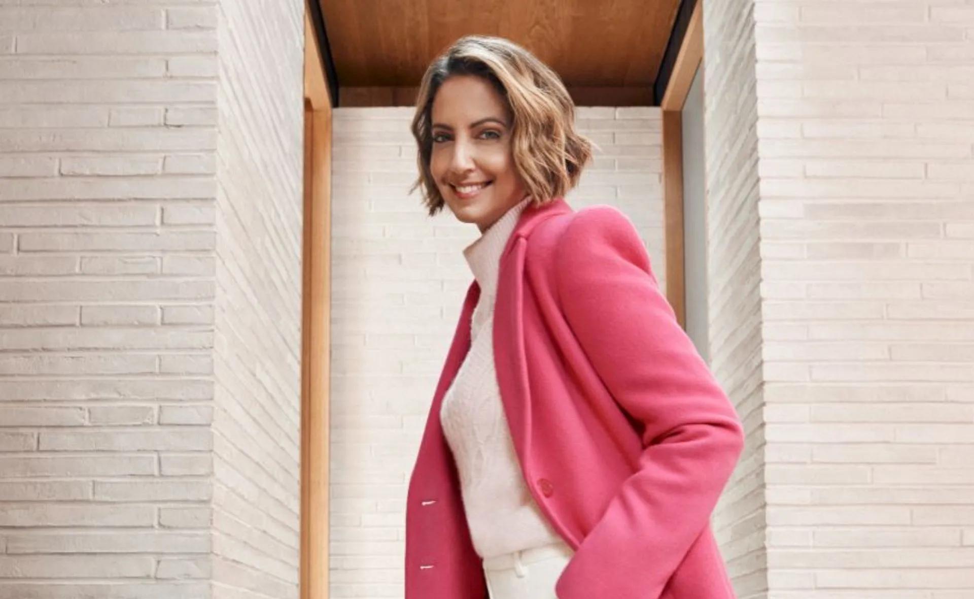 Smiling woman in a vibrant pink coat with a white sweater, standing by a brick wall.