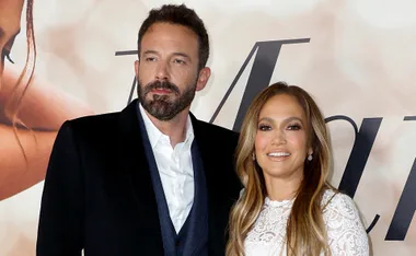 A couple on the red carpet, both dressed formally, smiling at an event backdrop.