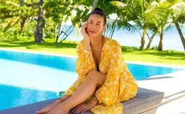 Woman in a yellow floral dress sits by a pool, surrounded by lush greenery and palm trees, smiling at the camera.