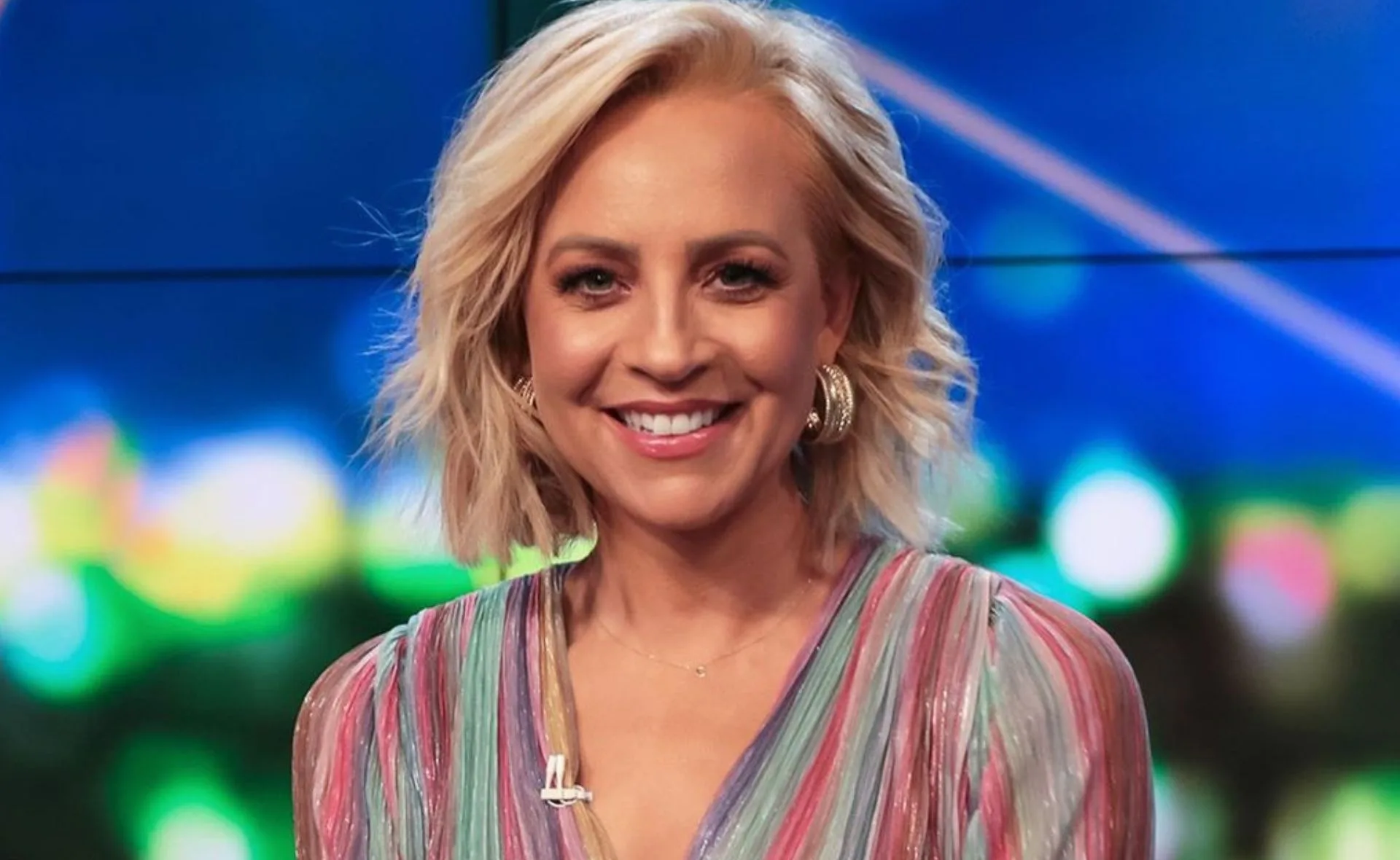 Smiling woman with short blonde hair, wearing a colorful striped top and hoop earrings, on a blurred colorful background.