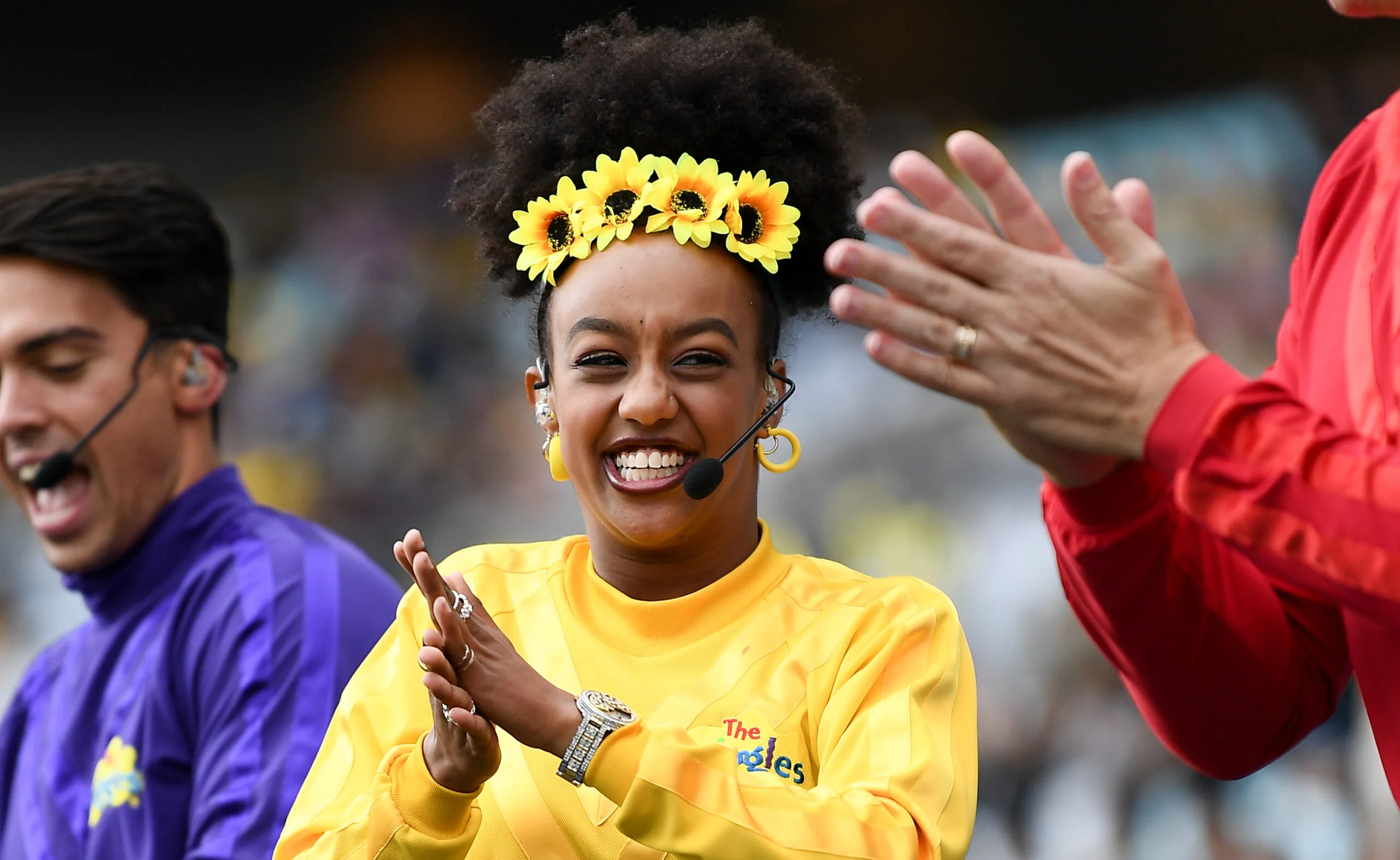 A person in a yellow shirt with a sunflower headband and headset, clapping joyfully with others in colorful outfits.