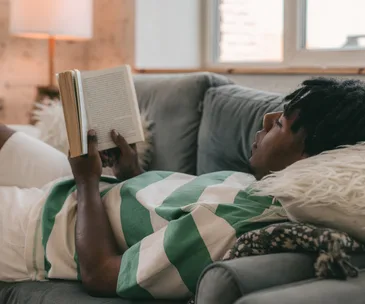 Man relaxing on a couch reading a book, with a lamp and window in the background.