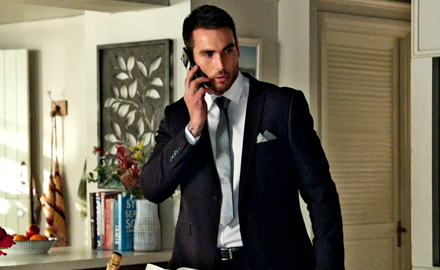 Man in a suit talking on a phone in a well-decorated room with books and plants in the background.