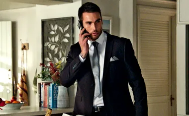 Man in a suit talking on a phone in a well-decorated room with books and plants in the background.