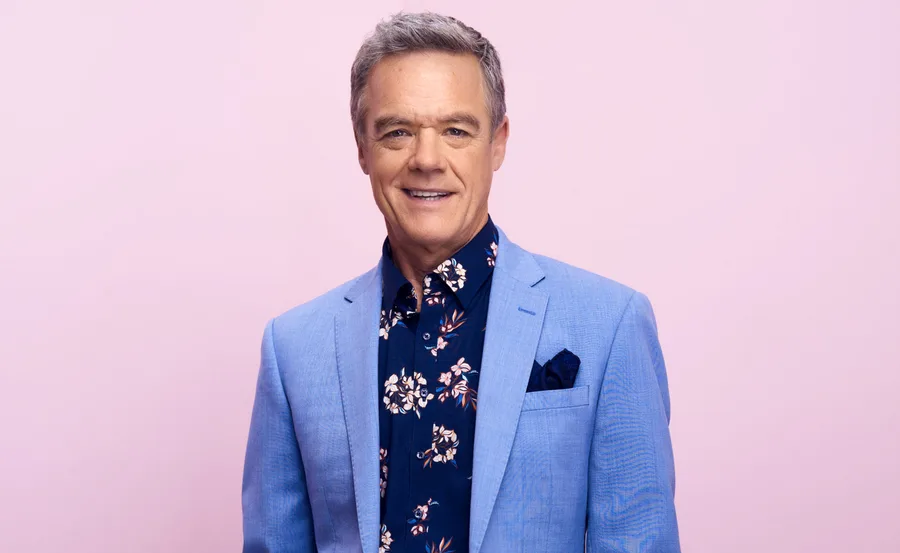 Man in a light blue suit and floral shirt against a pink background.