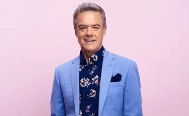 Man in a light blue suit and floral shirt against a pink background.