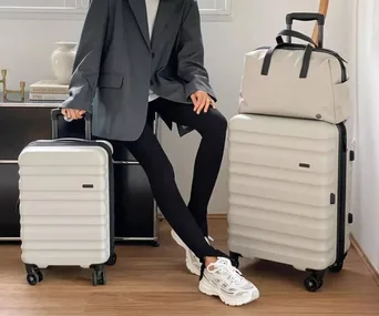 A person in a gray blazer sits, holding two cream-colored suitcases with wheels, on a wooden floor.