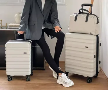 A person in a gray blazer sits, holding two cream-colored suitcases with wheels, on a wooden floor.