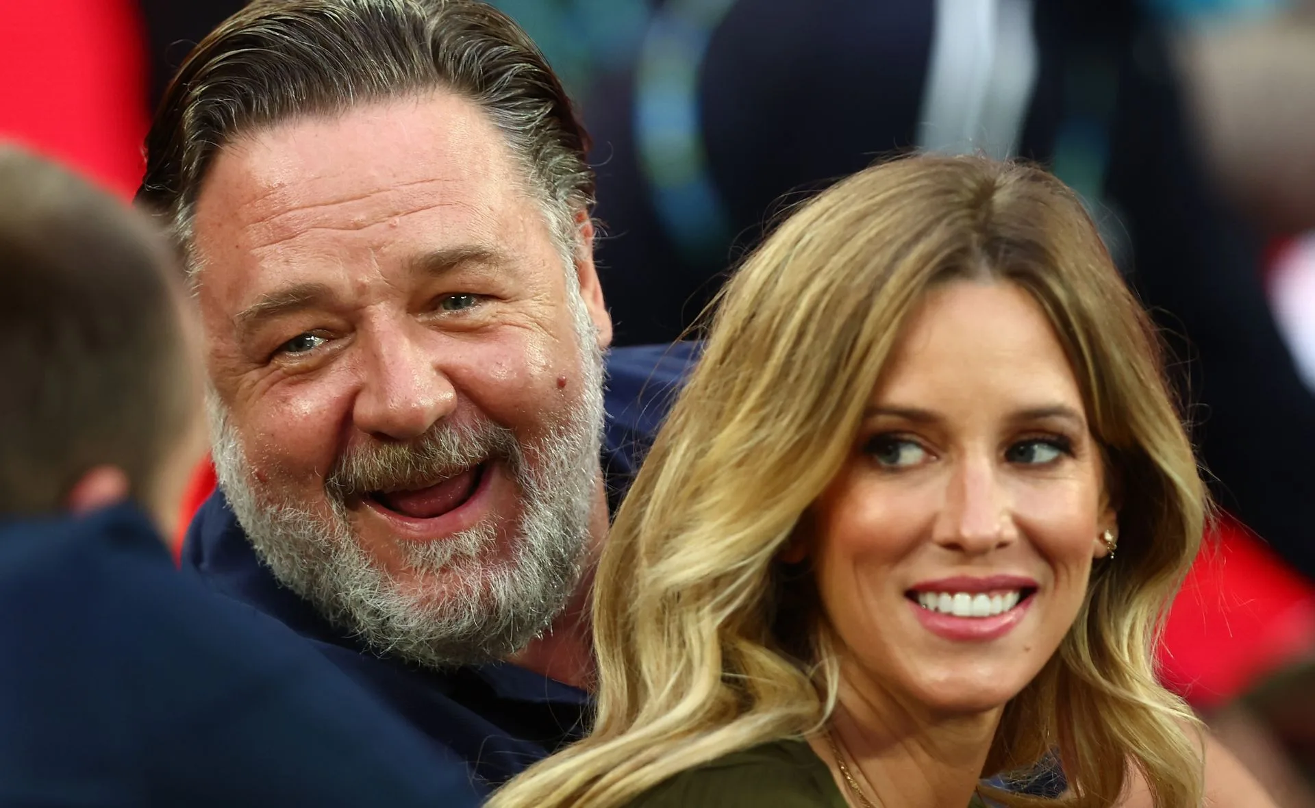 Man smiling joyfully with a woman who is looking away, both dressed casually outdoors.