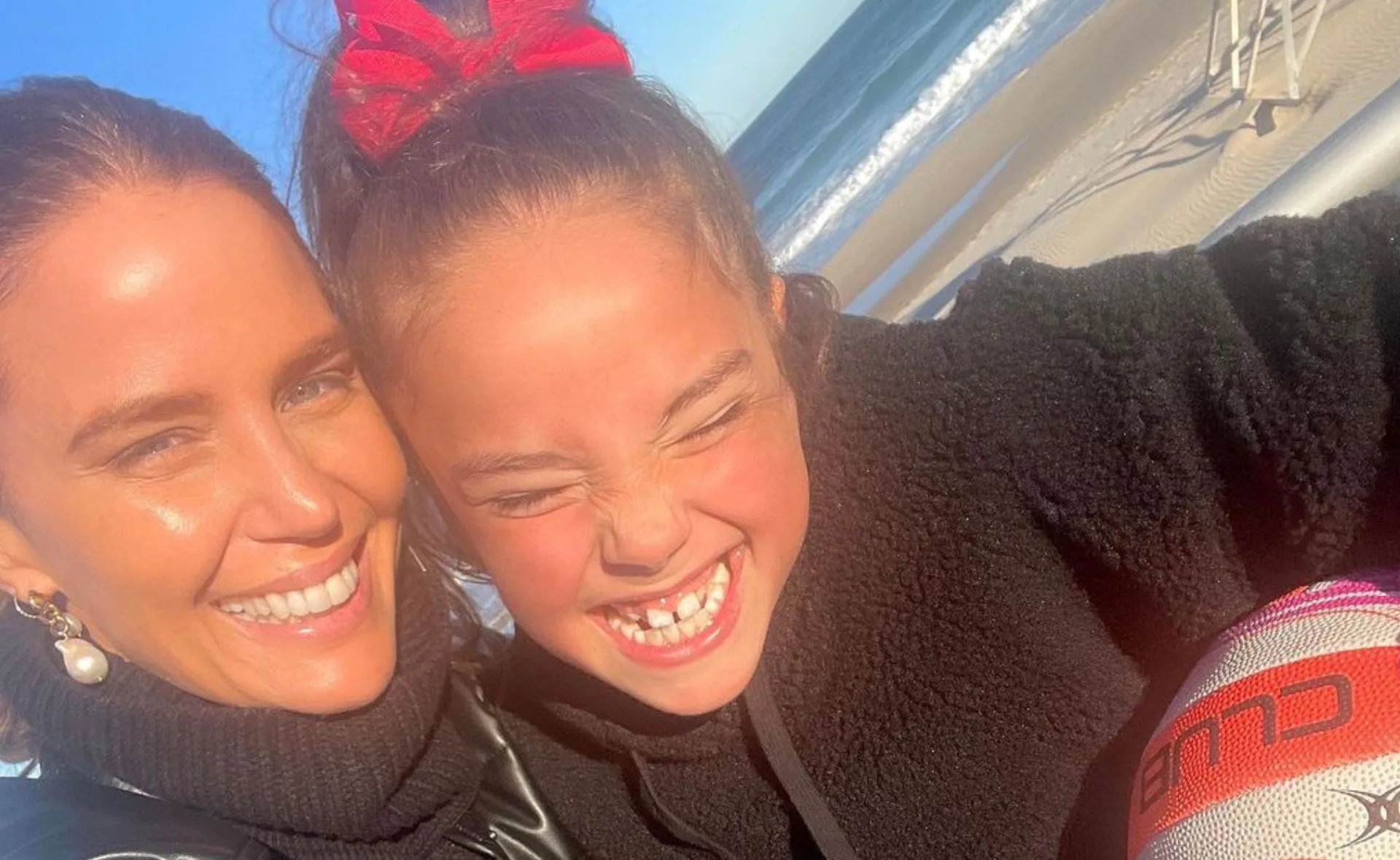 A woman and a young girl smile together at the beach, with the ocean in the background.