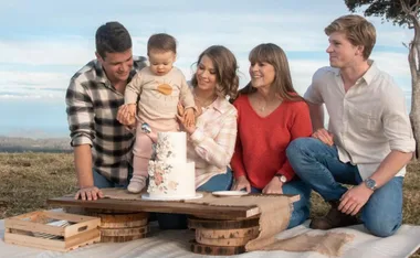 A family gathered outdoors with a toddler, sitting near a decorated birthday cake on a rustic table.