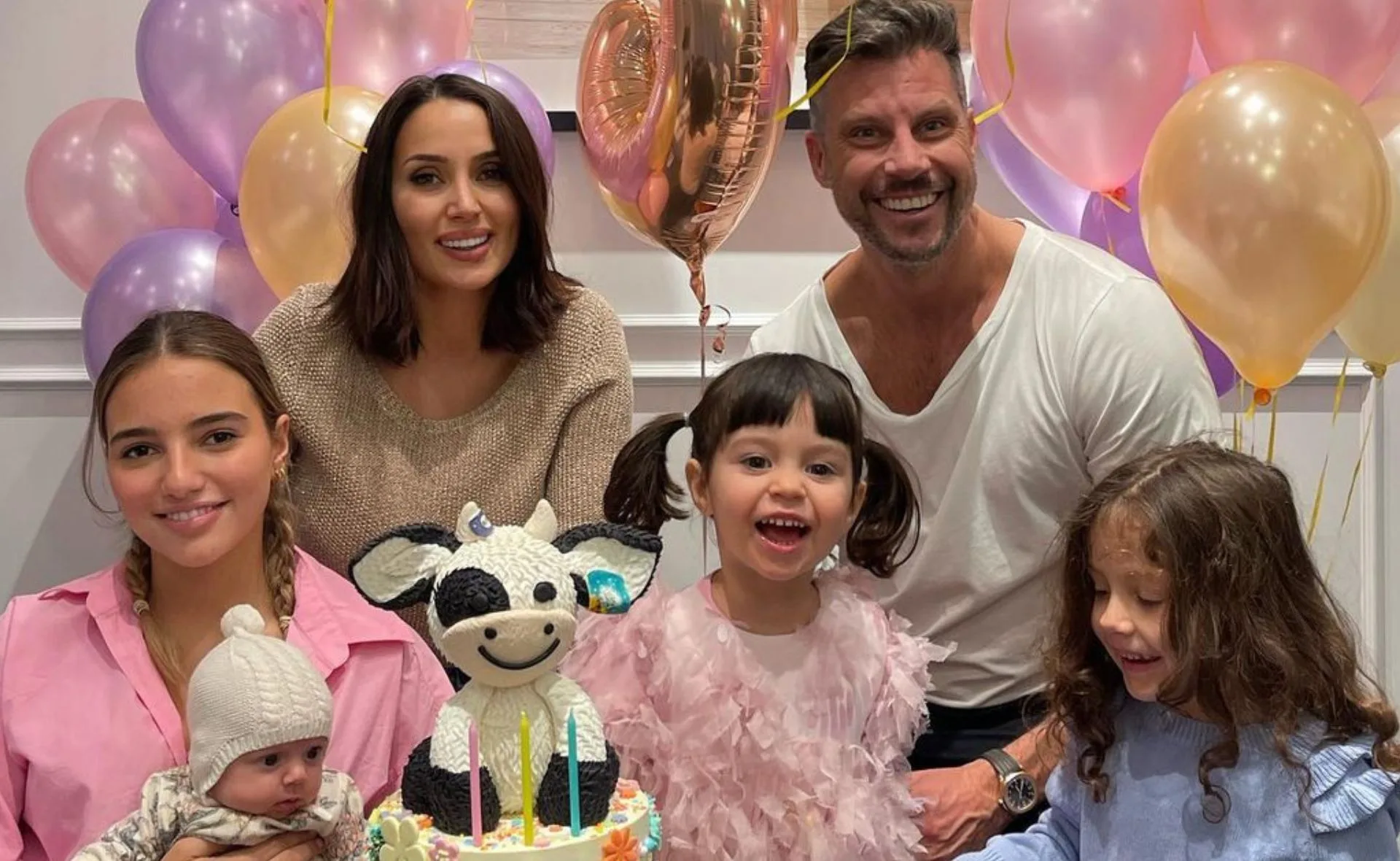 A family celebrates a child's birthday with balloons and a cow-themed cake, smiling happily together.