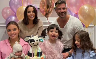 A family celebrates a child's birthday with balloons and a cow-themed cake, smiling happily together.
