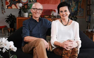 Elderly couple sitting on a sofa in a warmly decorated living room, both smiling at the camera.