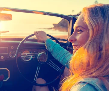 A woman driving a convertible near a beach during sunset, smiling and enjoying the scenic view.