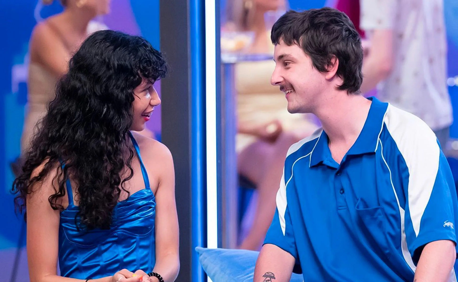 A woman in a blue dress and a man in a blue sports shirt smiling at each other in a vibrant room with people in the background.