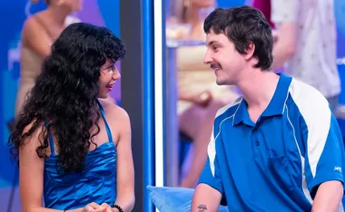 A woman in a blue dress and a man in a blue sports shirt smiling at each other in a vibrant room with people in the background.