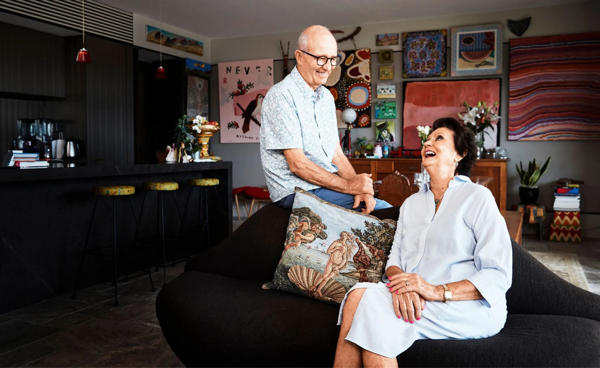 Elderly couple laughing together on a couch in a colorful, art-filled living room.