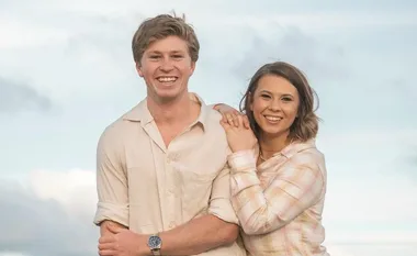 Two people smiling in light-colored shirts, standing close with arms around each other, against a cloudy sky backdrop.