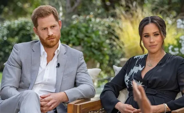 A man in a gray suit and a woman in a black dress sit outdoors, appearing in a formal interview setting.