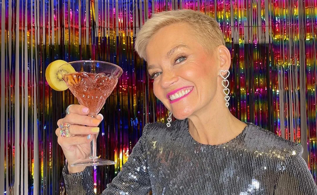 A smiling woman in a sparkly dress holds a cocktail with a kiwi slice, colorful tinsel backdrop.