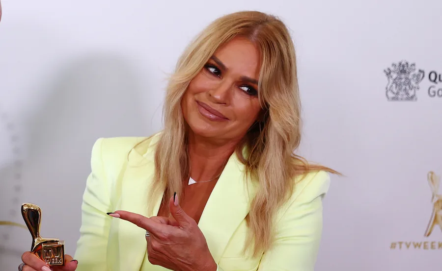 A woman in a yellow blazer pointing to a gold TV Week Logie award, smiling at an event backdrop.