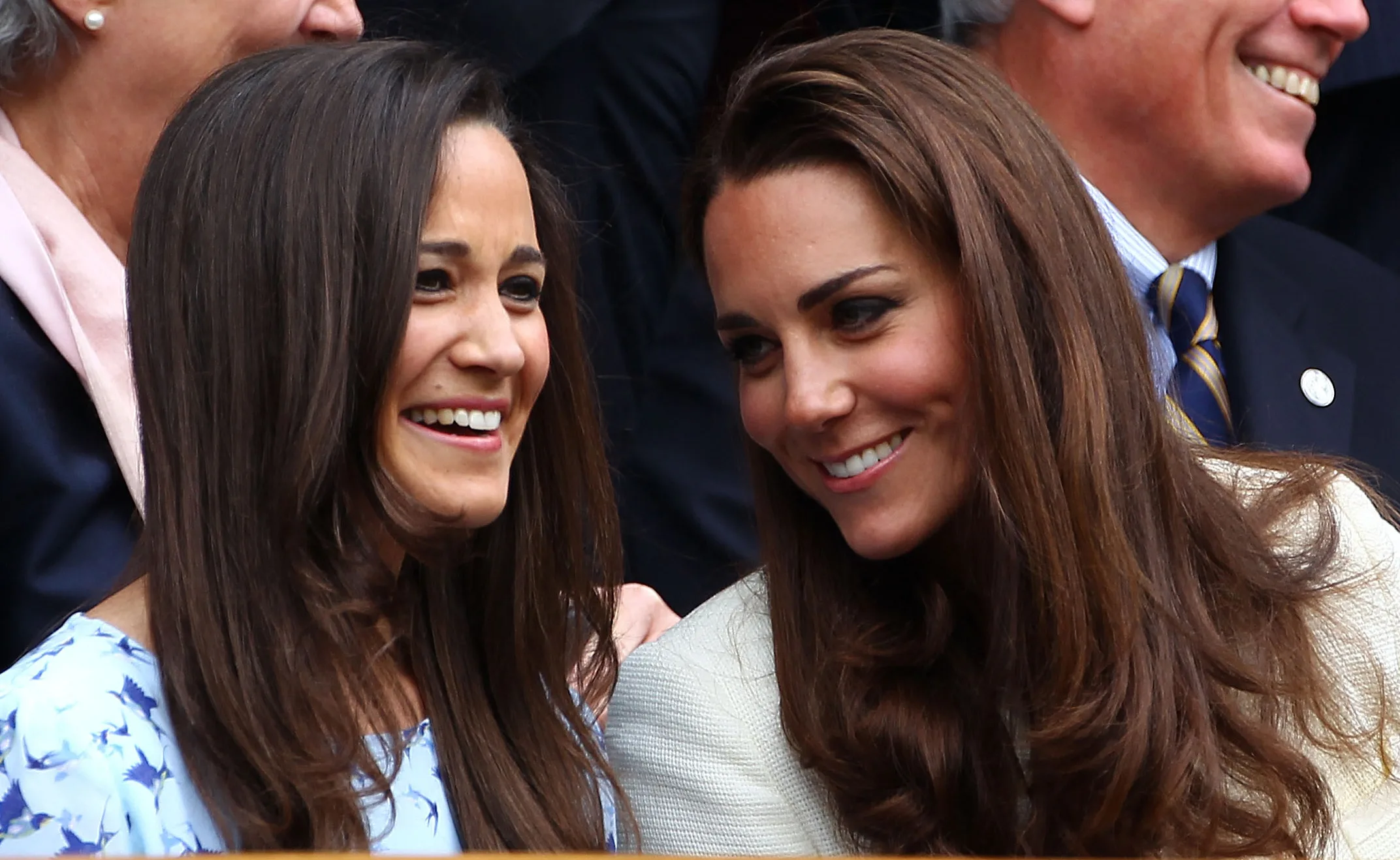 Two women with long brown hair laughing and smiling while seated, surrounded by others in formal attire.