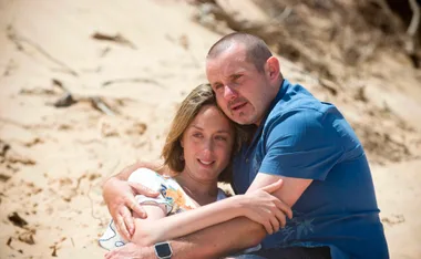A couple embracing on a sandy beach, both with content expressions.