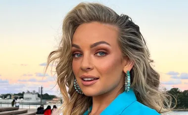 A person with wavy blonde hair wearing turquoise earrings and a blue outfit, with a sunset background.