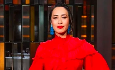 A woman in a red dress with blue earrings stands confidently in front of a patterned backdrop.