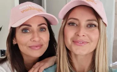 Two women smiling, wearing pink baseball caps with text, indoors.