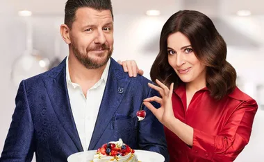 A man in a blue suit holds a dessert while a woman in a red shirt decorates it with a cherry.