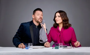 Man and woman sitting at a table; man in blue suit, woman in pink blouse playfully wipes his face with napkin.