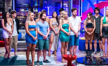 Contestants standing together on a reality show set with blue and pink lighting.